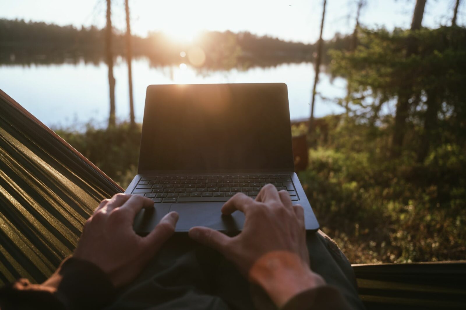 Henkilö istuu riippukeinussa järven rannalla kannettava tietokone sylissään